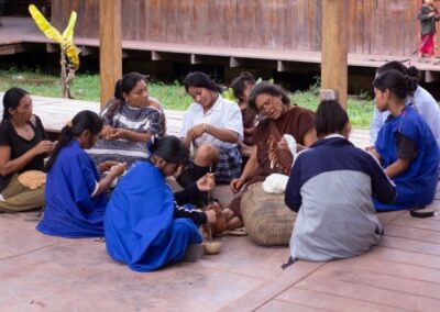 Gruppe von indigenen Frauen und Mädchen sitzen im Kreis auf einer Holzveranda und spinnen, bzw. weben Wolle
