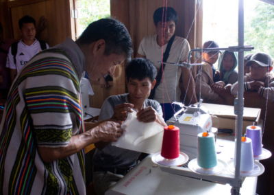 Zwei Männer arbeiten an einer Nähmaschine mit bunten Garnrollen, während Kinder und junge Erwachsene aufmerksam zuschauen